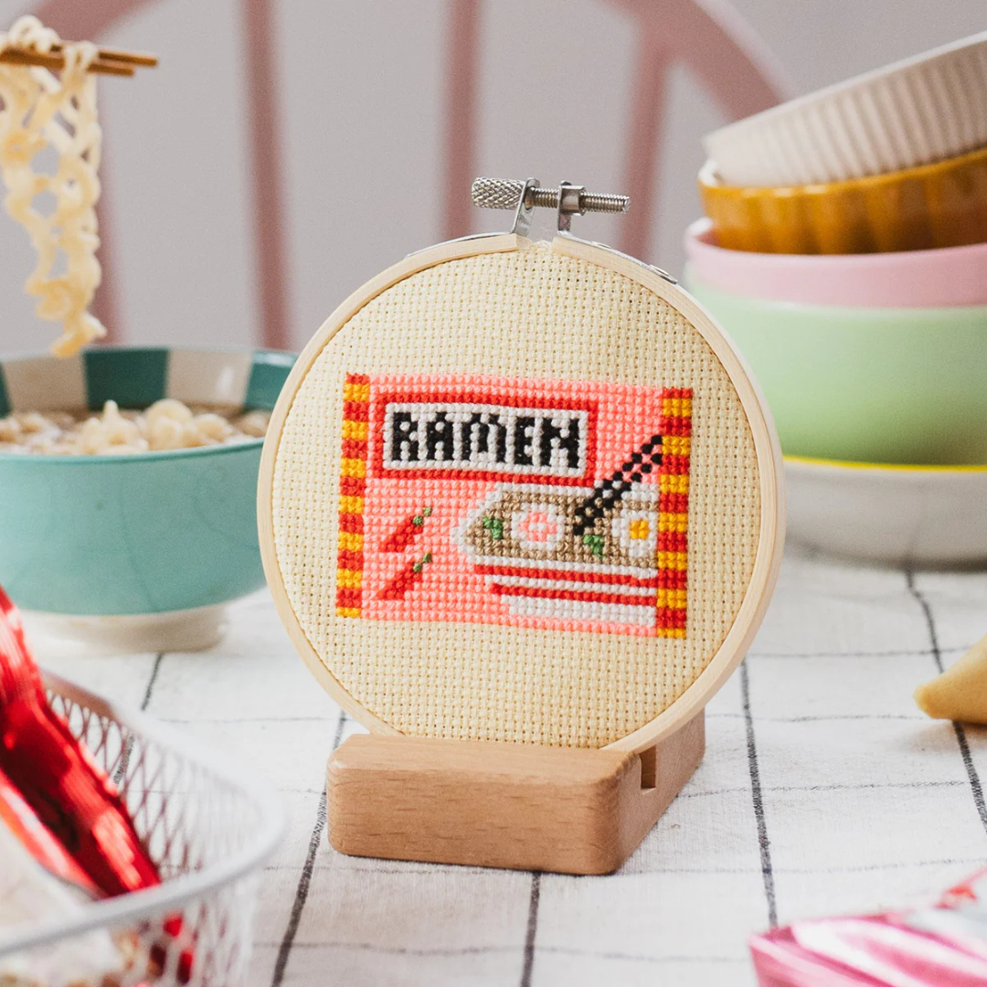 Finished ramen cross stitch displayed in a bamboo embroidery hoop on a wooden stand.