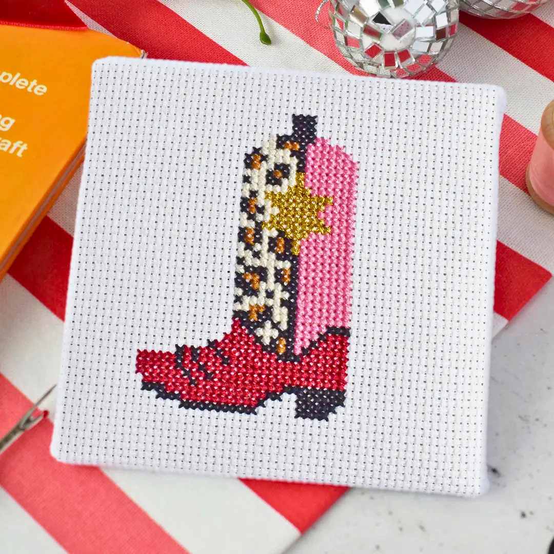 A square white cross-stitch canvas featuring a cowboy boot design in red, pink, black, white, and gold, styled on a red and white striped surface with craft props.