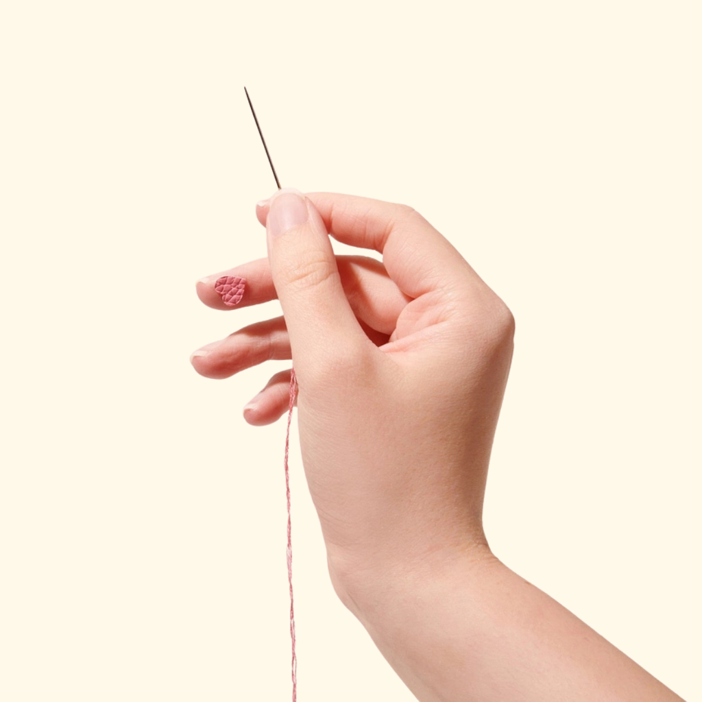 Close-up of a hand holding a sewing needle, demonstrating the use of SOHMO Adhesive Thimble Pads for hand sewing and embroidery.