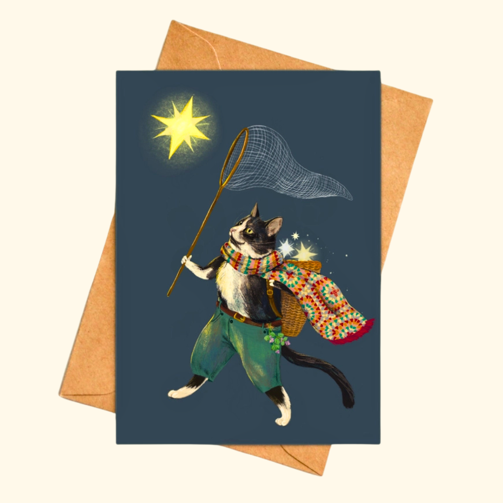 Ashy Cat greeting card shown with a matching kraft envelope, featuring a whimsical cat wearing a flowing cape and a basket on his back while trying to catch glowing stars with a fishing net.