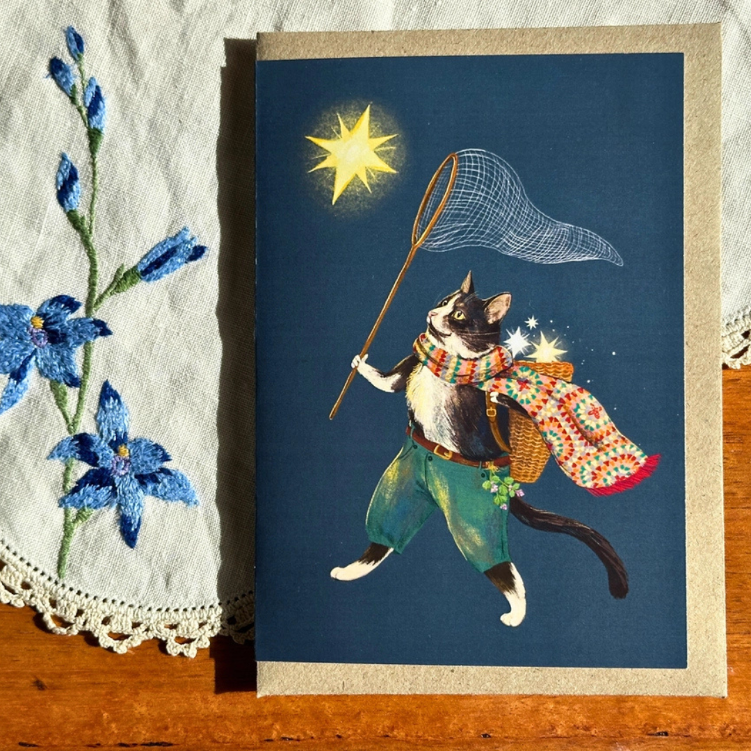 Ashy Cat greeting card styled on a wooden surface with lace and blue flowers, featuring a whimsical cat wearing a patterned cape and carrying a basket of glowing stars while catching another with a fishing net.
