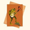Banjo Frog greeting card shown with a matching kraft envelope, featuring a cheerful green frog playing a banjo while a butterfly perches on the instrument.
