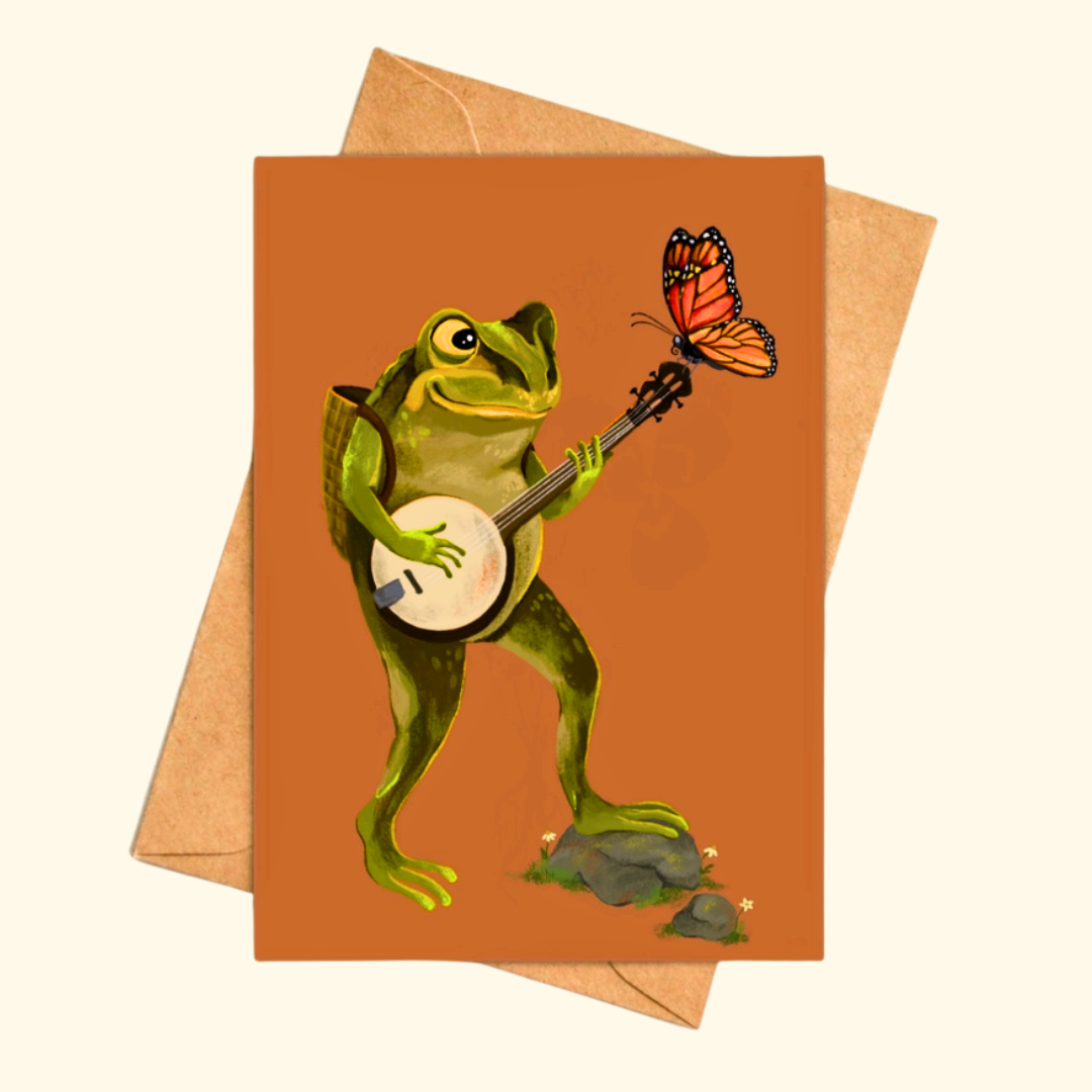 Banjo Frog greeting card shown with a matching kraft envelope, featuring a cheerful green frog playing a banjo while a butterfly perches on the instrument.