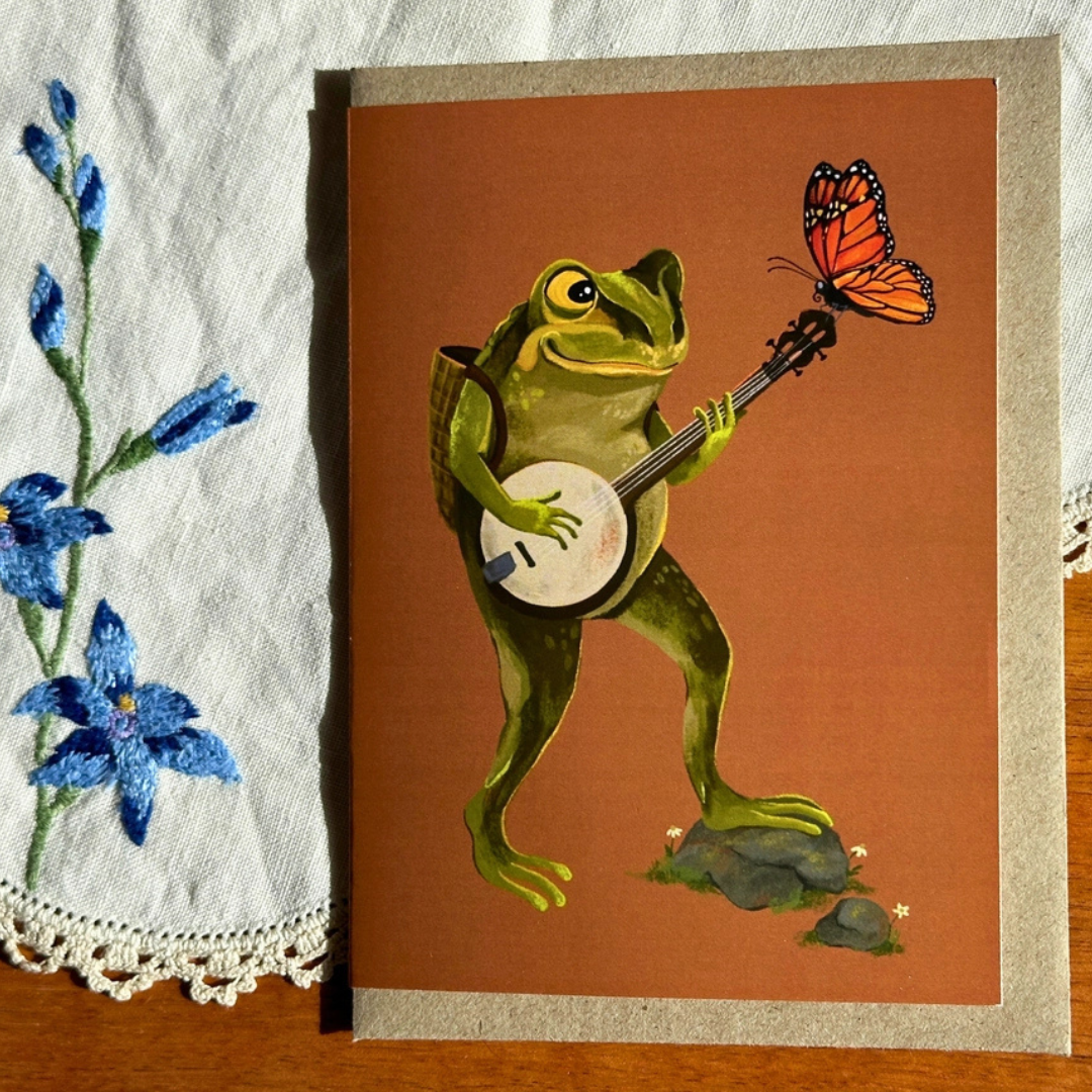 Banjo Frog greeting card styled on a wooden surface with lace and blue flowers, featuring a charming green frog playing a banjo as a butterfly perches on the instrument.