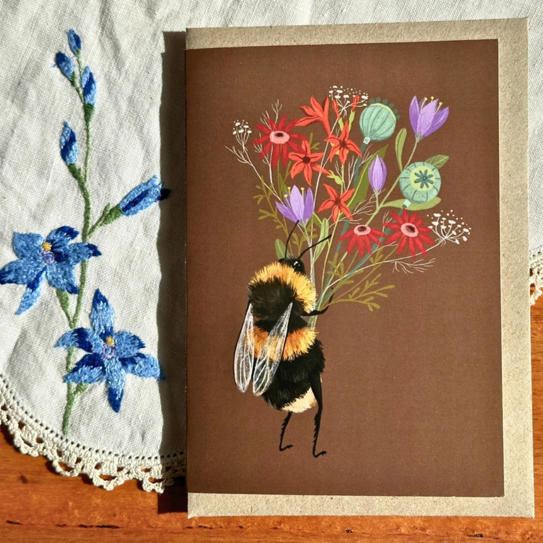 Bee With A Posy greeting card styled on a wooden surface with lace and blue flowers, featuring a charming bumblebee carrying a vibrant bouquet of wildflowers.
