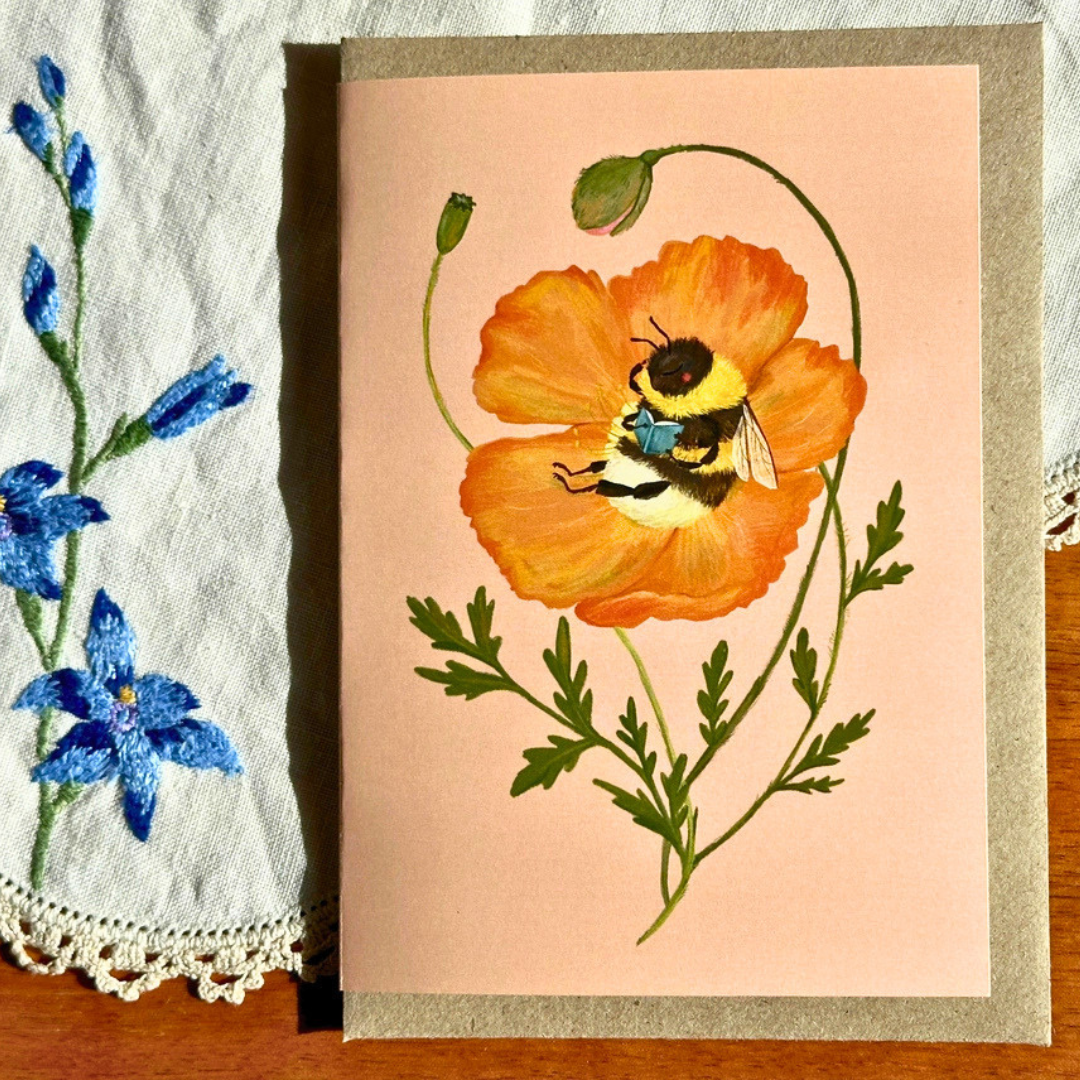 Beebliophile greeting card styled on a wooden surface with lace and blue flowers, featuring a delightful bee reading a book while nestled inside a vibrant orange poppy.