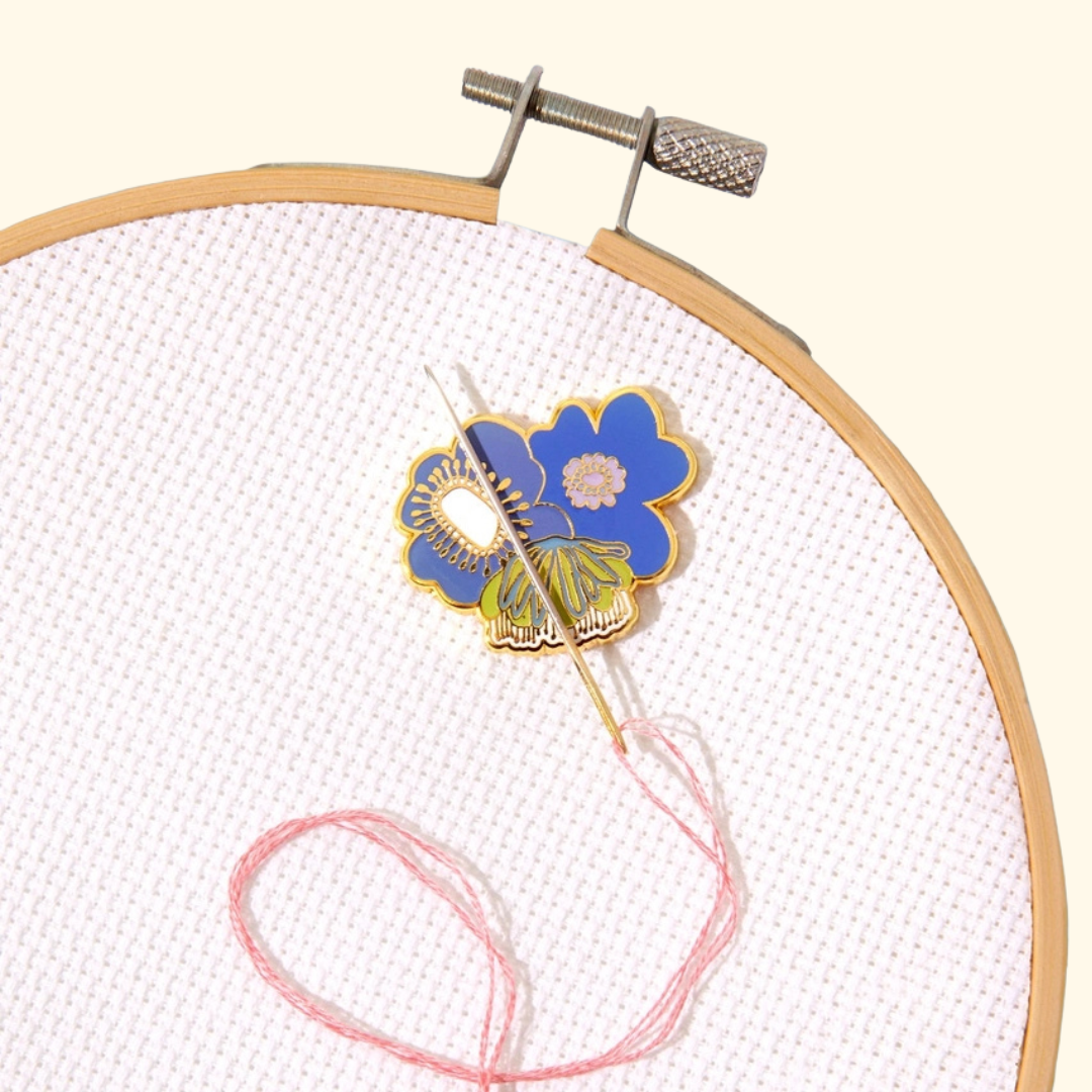 A close-up of a floral enamel pin with blue, yellow, and green details, placed on white cross-stitch fabric inside a wooden embroidery hoop with pink thread.