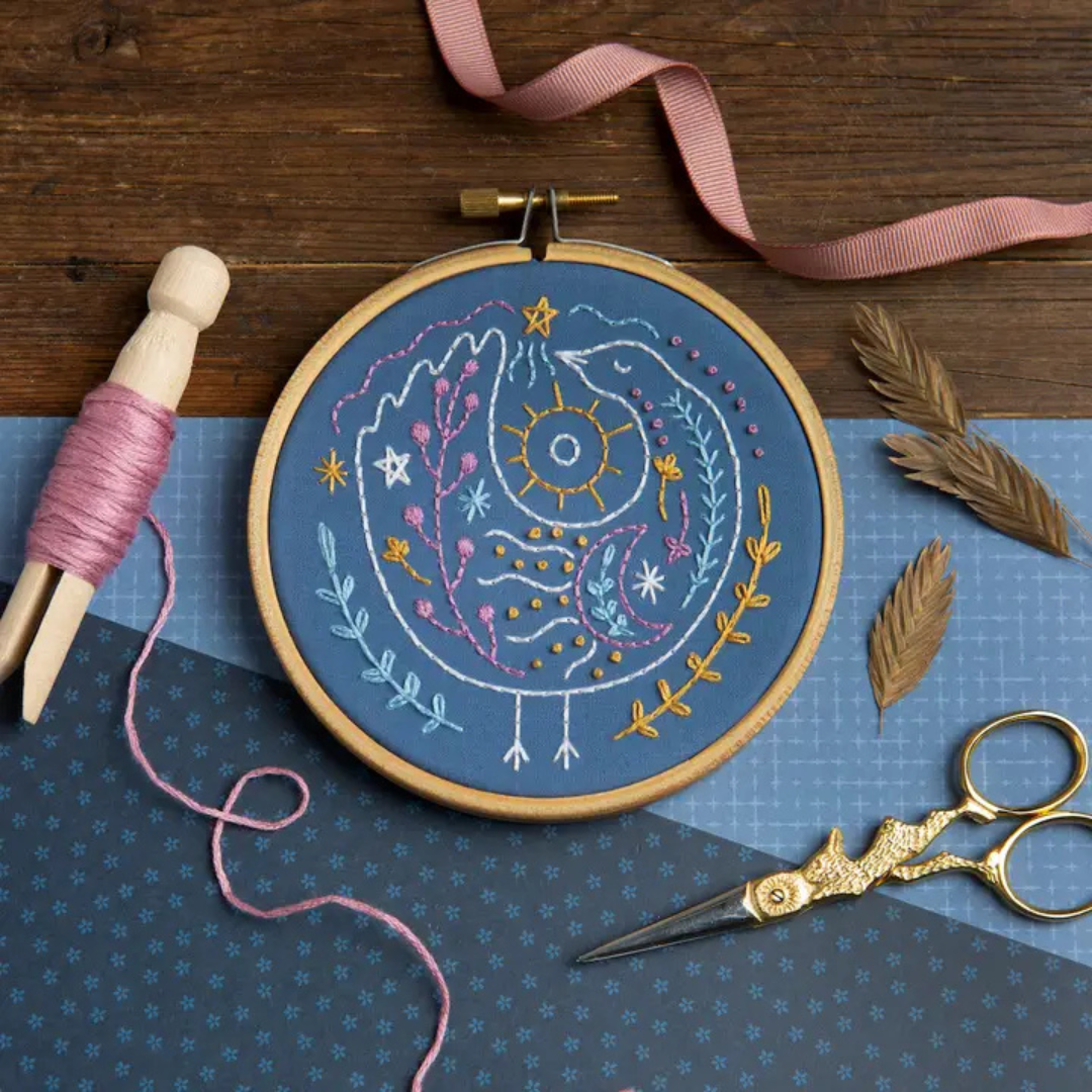 Celestial Bird Mini Embroidery Kit in progress with blue fabric, wooden embroidery hoop, embroidery thread and scissors.