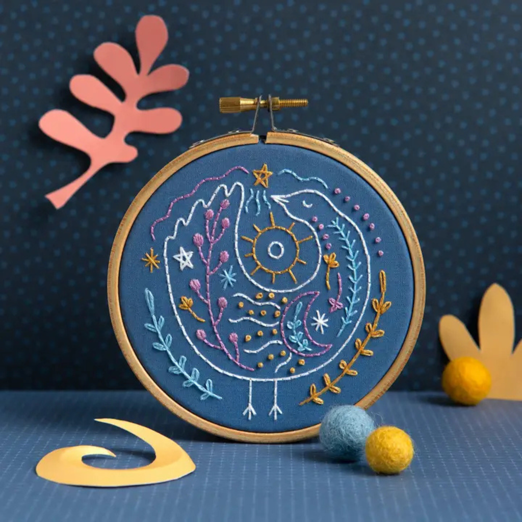 Finished Celestial Bird Mini Embroidery Kit displayed in a wooden hoop against a dark blue background with colourful craft decorations.