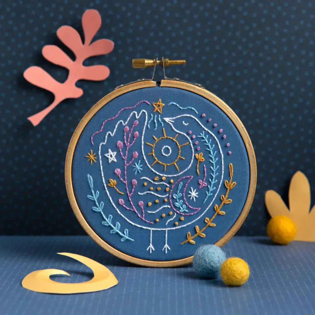 Finished Celestial Bird Mini Embroidery Kit displayed in a wooden hoop against a dark blue background with colourful craft decorations.