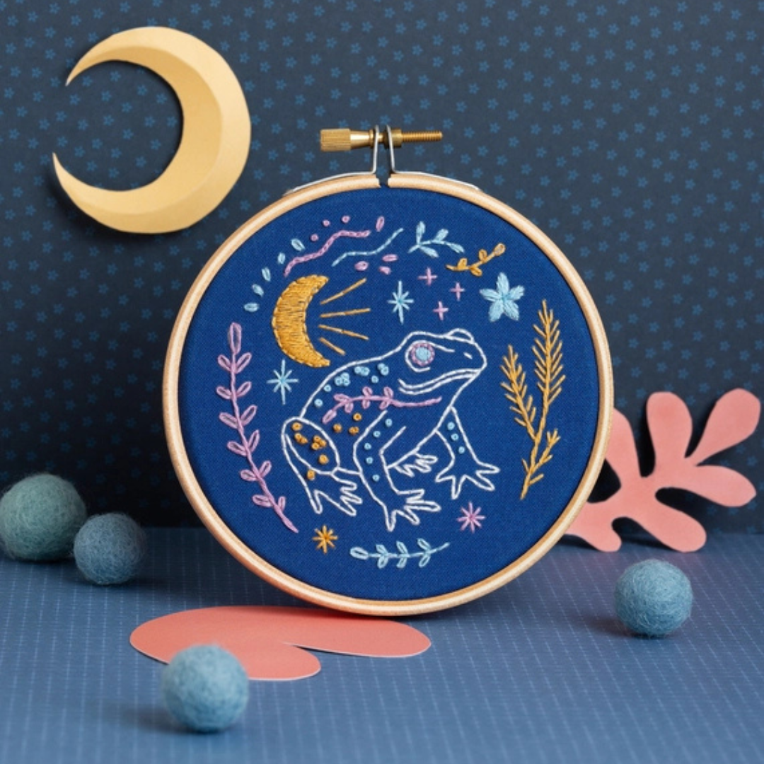 Finished Celestial Frog embroidery hoop displayed with decorative moon and coral props.