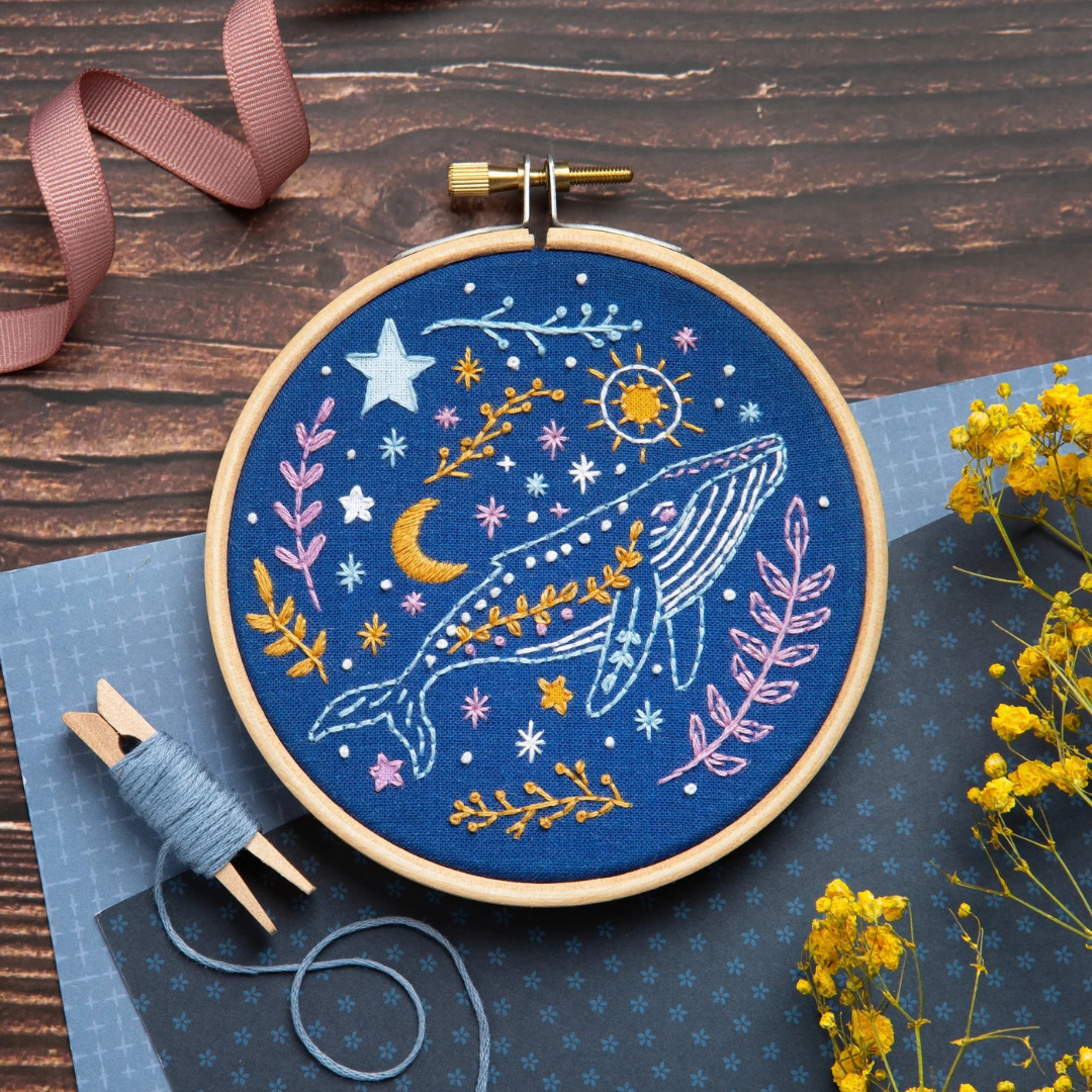 Celestial whale embroidery hoop displayed in a flat lay with embroidery thread, ribbon and floral styling elements.