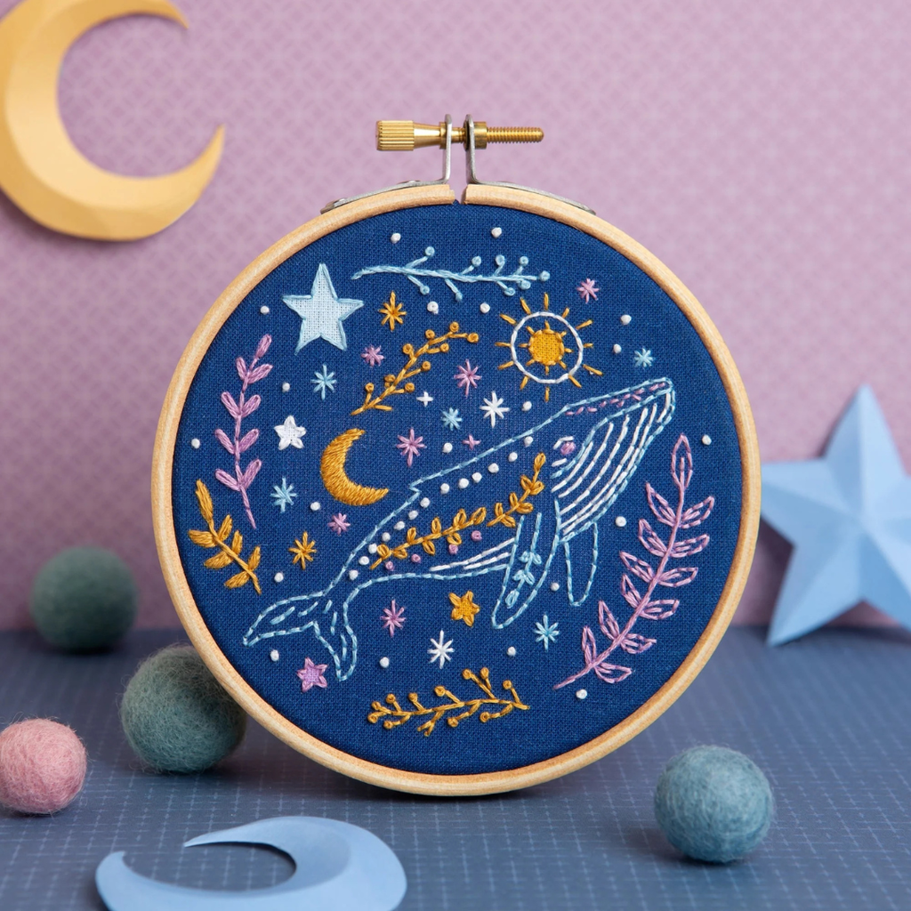 Finished celestial whale embroidery design displayed upright with decorative stars, pom poms and moon themed props.