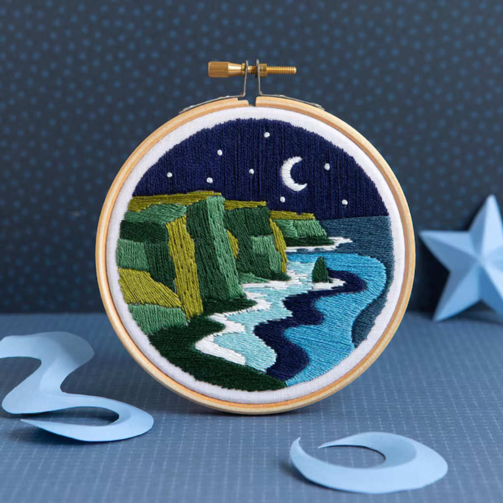 Finished Celtic Cliffs embroidery project in a wooden hoop featuring dramatic Irish coastline scenery under a starry night sky.