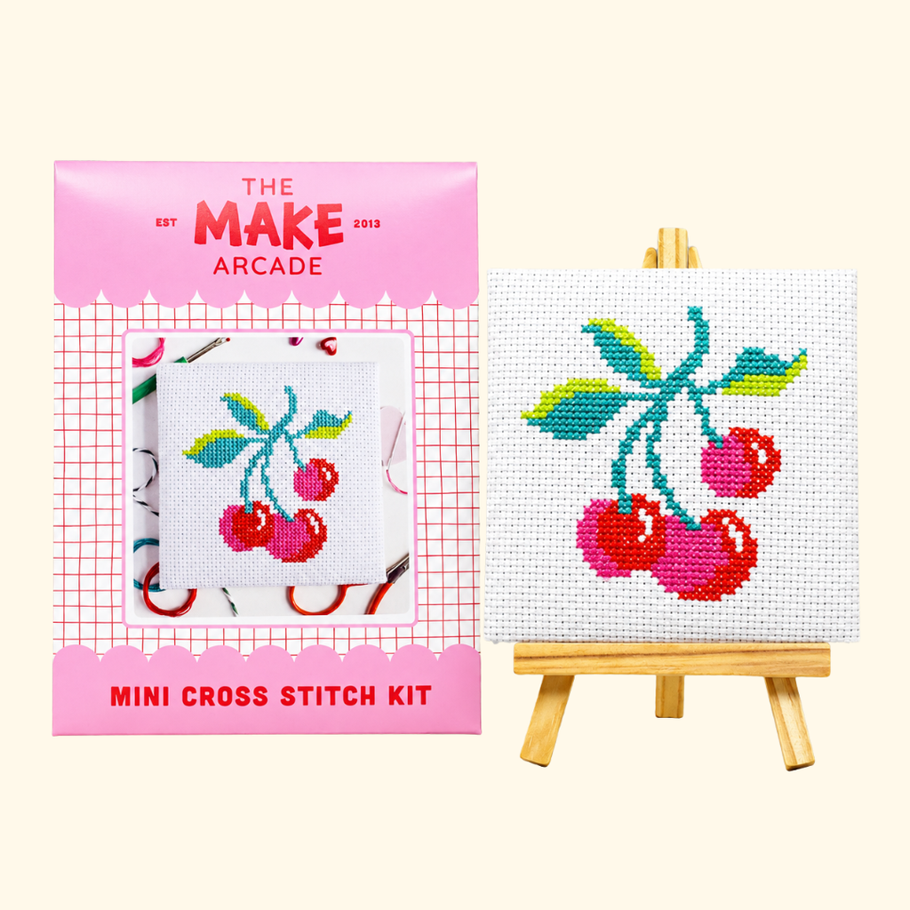 The Make Arcade Cherries Mini Cross Stitch Kit beside the completed design on a miniature easel.