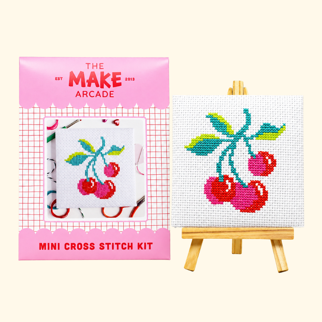 The Make Arcade Cherries Mini Cross Stitch Kit beside the completed design on a miniature easel.