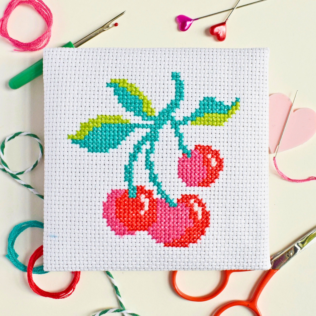 Cherries mini cross stitch project surrounded by embroidery threads, scissors and craft tools.