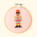 Finished Christmas nutcracker cross stitch in colourful thread, displayed in a bamboo embroidery hoop on pink Aida fabric.