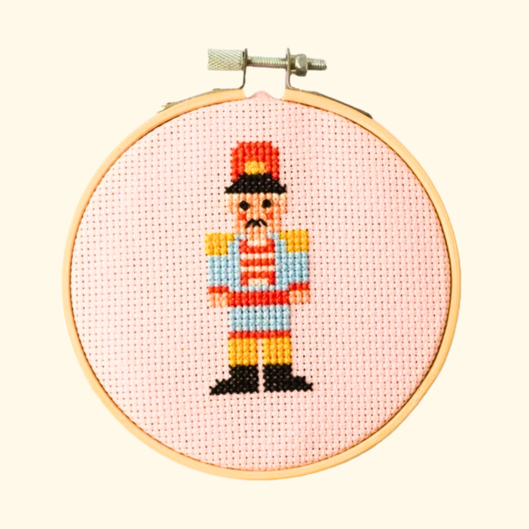 Finished Christmas nutcracker cross stitch in colourful thread, displayed in a bamboo embroidery hoop on pink Aida fabric.