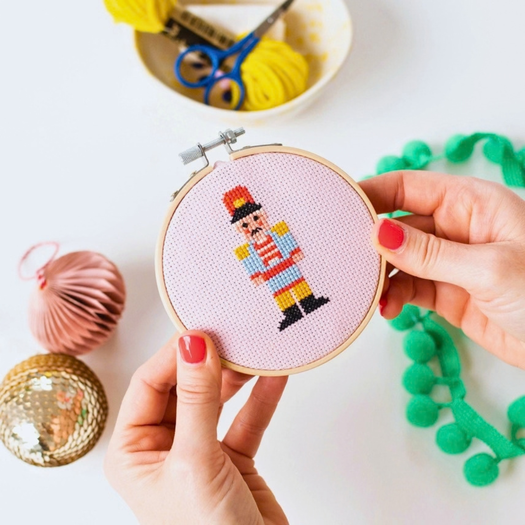 Christmas nutcracker cross stitch being held in a bamboo hoop, surrounded by colourful craft supplies.