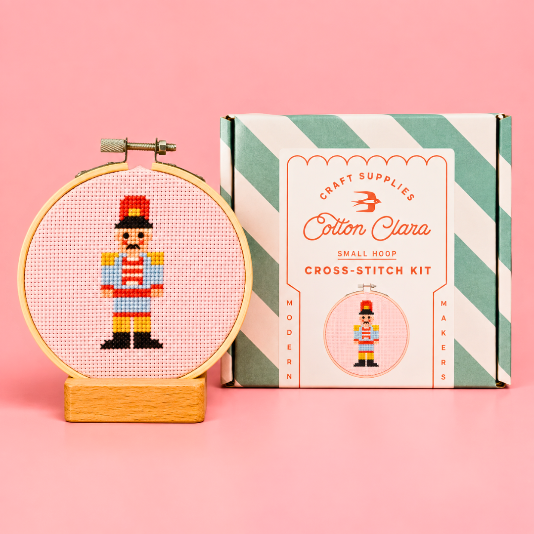 Completed Christmas nutcracker cross stitch in a bamboo hoop displayed beside the Cotton Clara kit box on a pink background.