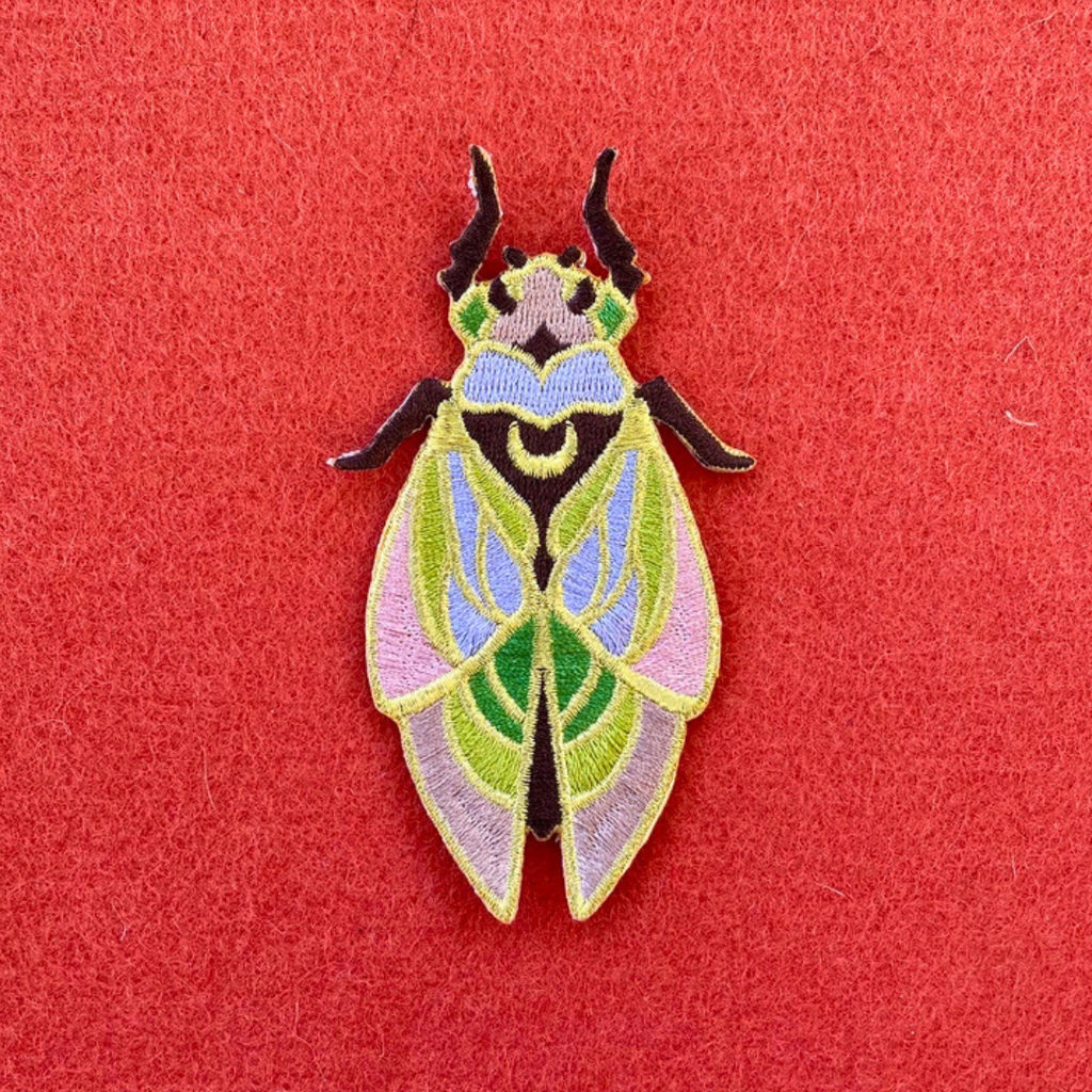 Close-up of Ashlea Bechaz Illustration cicada embroidered iron on patch featuring intricate green and pink wings, colourful body detailing and dark antennae against a coral red background.
