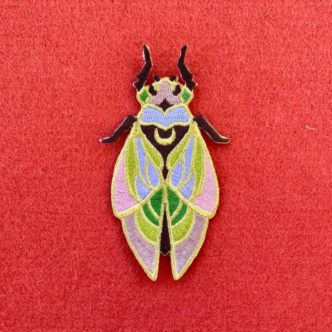 Close-up of Ashlea Bechaz Illustration cicada embroidered iron on patch featuring intricate green and pink wings, colourful body detailing and dark antennae against a coral red background.