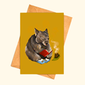 Cosy Wombat greeting card shown with matching kraft envelope, featuring an illustrated wombat in slippers reading a red book beside a steaming cup of tea.