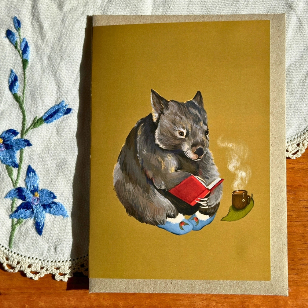 Cosy Wombat greeting card styled on a wooden surface with lace and blue flowers, featuring an illustrated wombat reading a book with a cup of tea nearby.