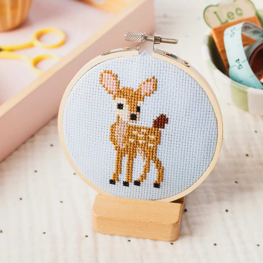 Finished deer cross stitch displayed in an embroidery hoop on a wooden stand.