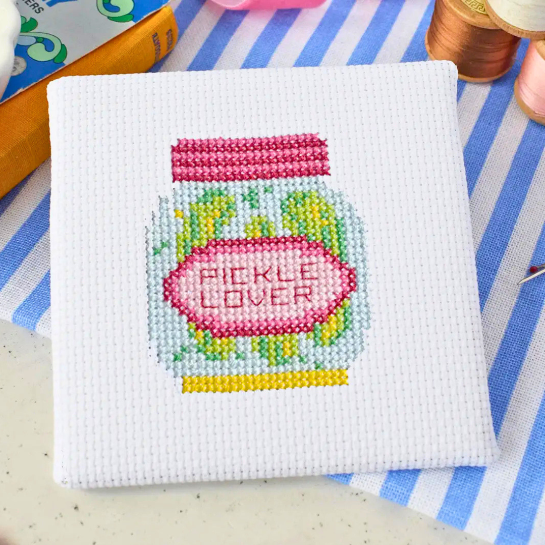 Finished Dill Pickle Mini Cross Stitch Kit displayed with embroidery scissors, thread and colourful craft supplies.