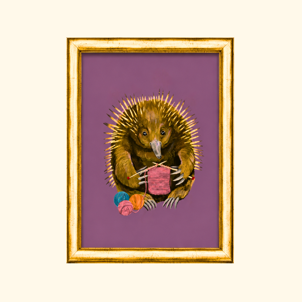 Echidna Knitter greeting card displayed in a gold frame, showcasing the whimsical illustration of an echidna knitting a pink scarf with colourful yarn.