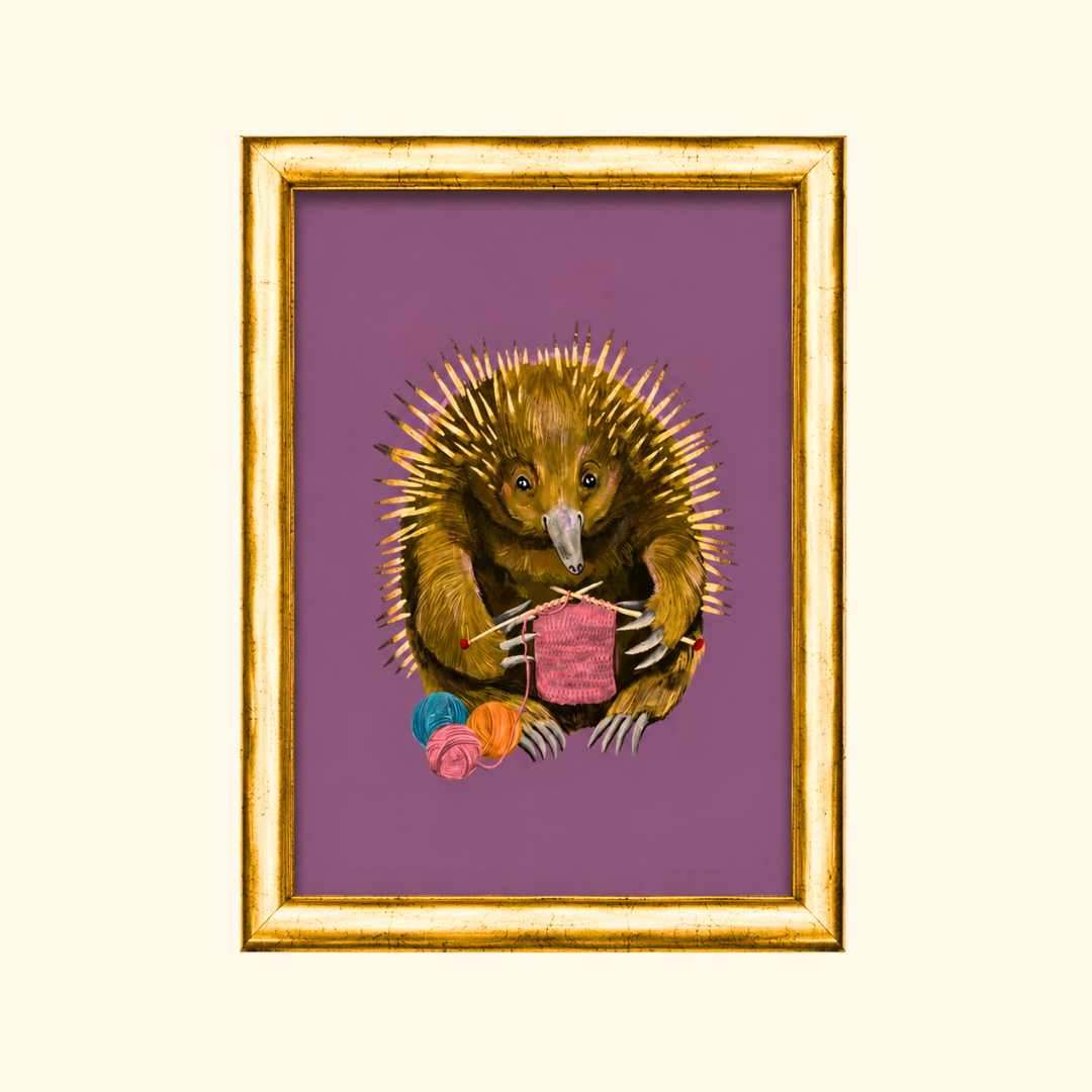 Echidna Knitter greeting card displayed in a gold frame, showcasing the whimsical illustration of an echidna knitting a pink scarf with colourful yarn.