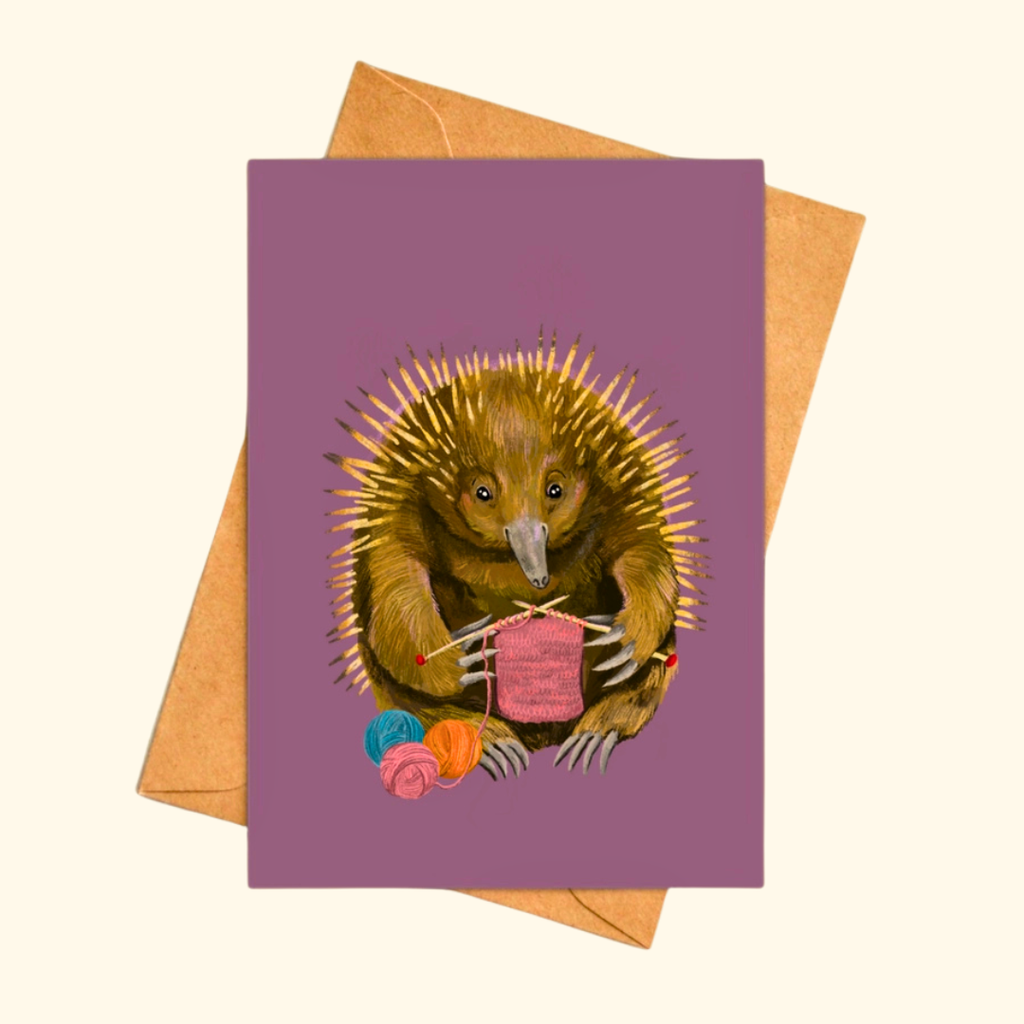 Echidna Knitter greeting card shown with a matching kraft envelope, featuring a charming echidna knitting a pink scarf surrounded by colourful balls of yarn.
