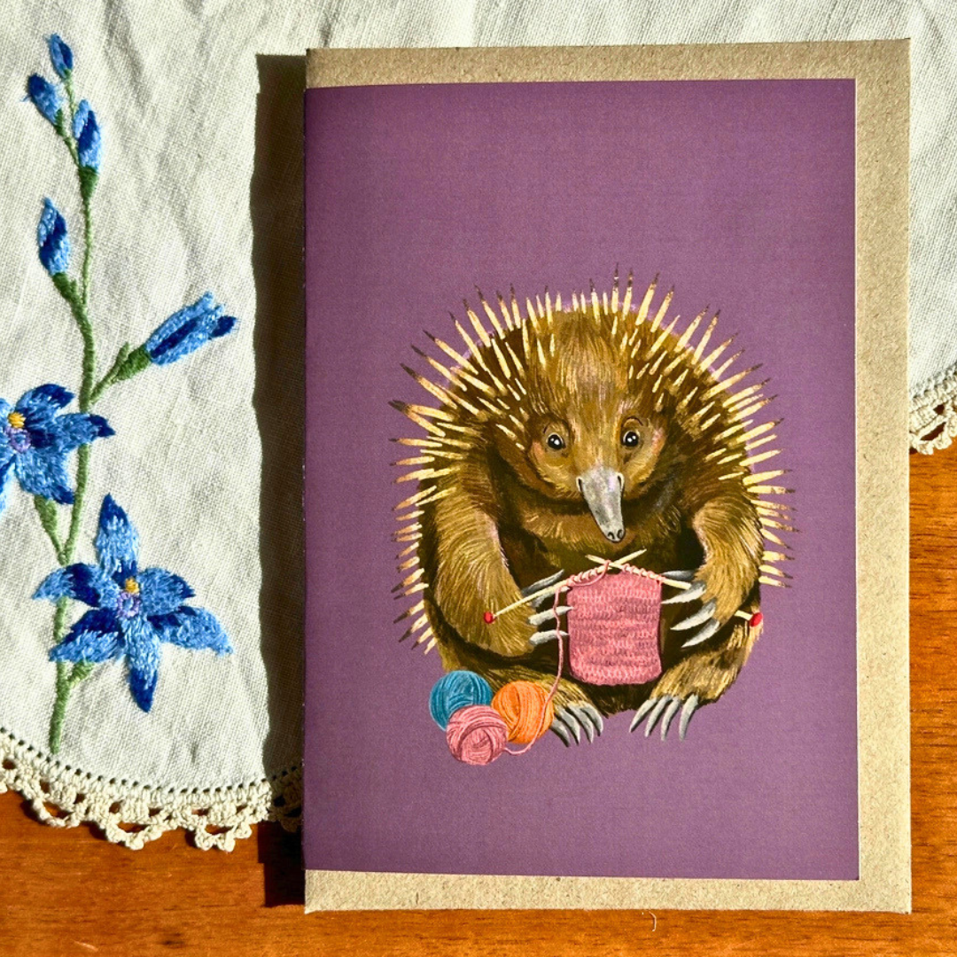Echidna Knitter greeting card styled on a wooden surface with lace and blue flowers, featuring a delightful echidna knitting a pink scarf surrounded by colourful balls of yarn.