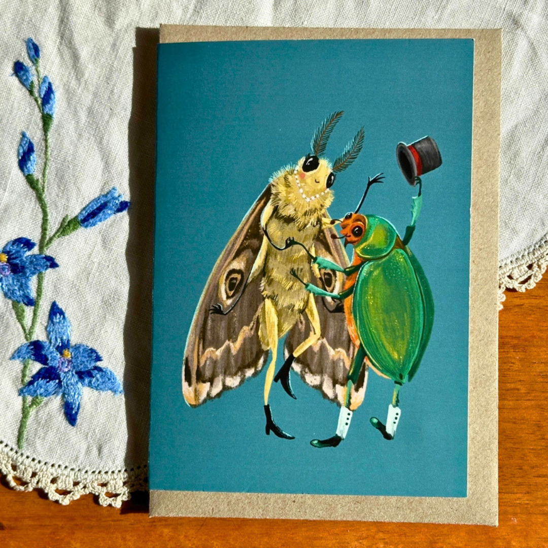 Fancy Dancing greeting card styled on a wooden surface with lace and blue flowers, featuring a whimsical moth lady and beetle gent enjoying a graceful dance together.