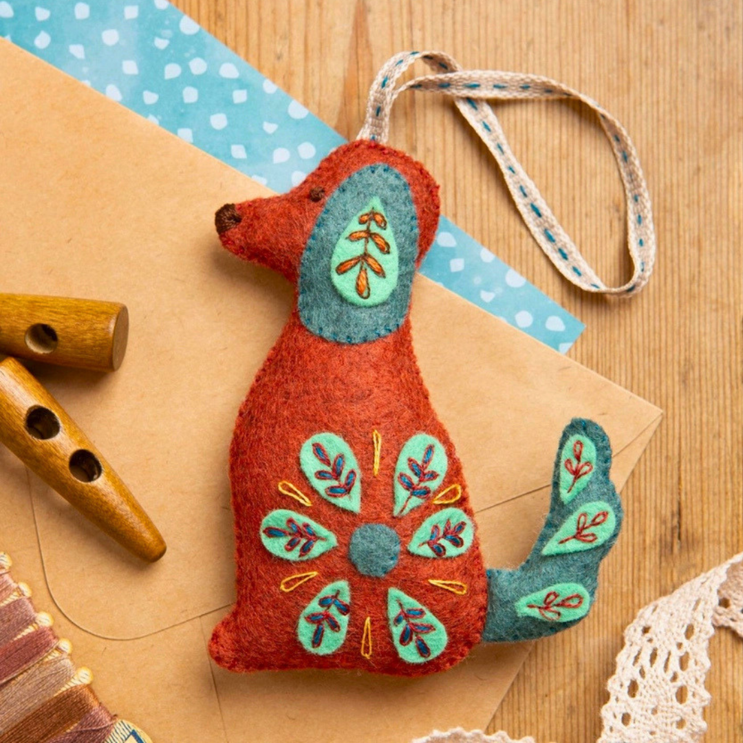 Completed Folk Dog felt ornament displayed with craft materials, showing its embroidered body, tail and hanging loop.