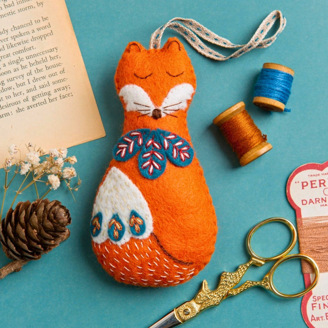 Completed Folk Fox felt ornament displayed with embroidery thread, scissors, pine cone and craft materials.