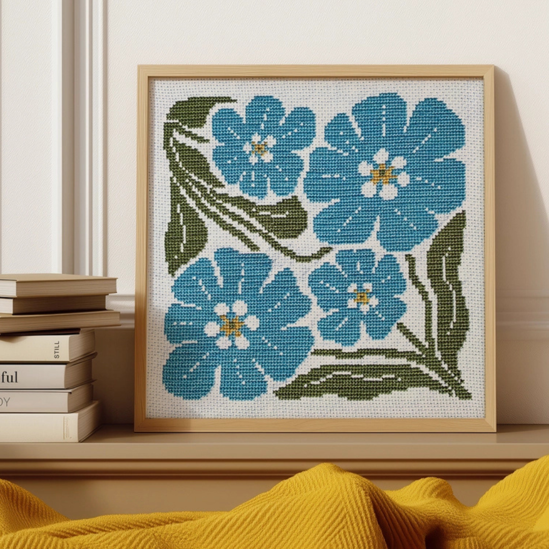 Framed Forget Me Not Grid cross stitch design by Budgie Goods displayed on a shelf with books and home decor.