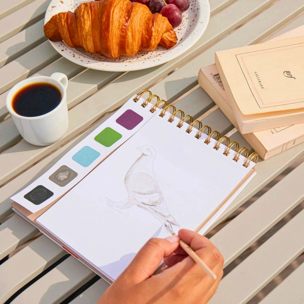 France Watercolour Travel Book by La Petite Epicerie being used at an outdoor café table, with a paintbrush, watercolour palette and croissant.