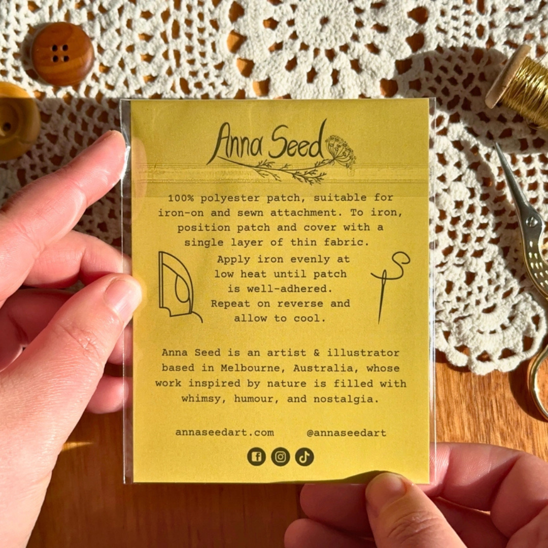 A person's hands hold a clear plastic sleeve containing a yellow backing card with instructions for an Anna Seed polyester patch. The card features black text and simple line drawings of an iron, a patch, and a sewing needle.