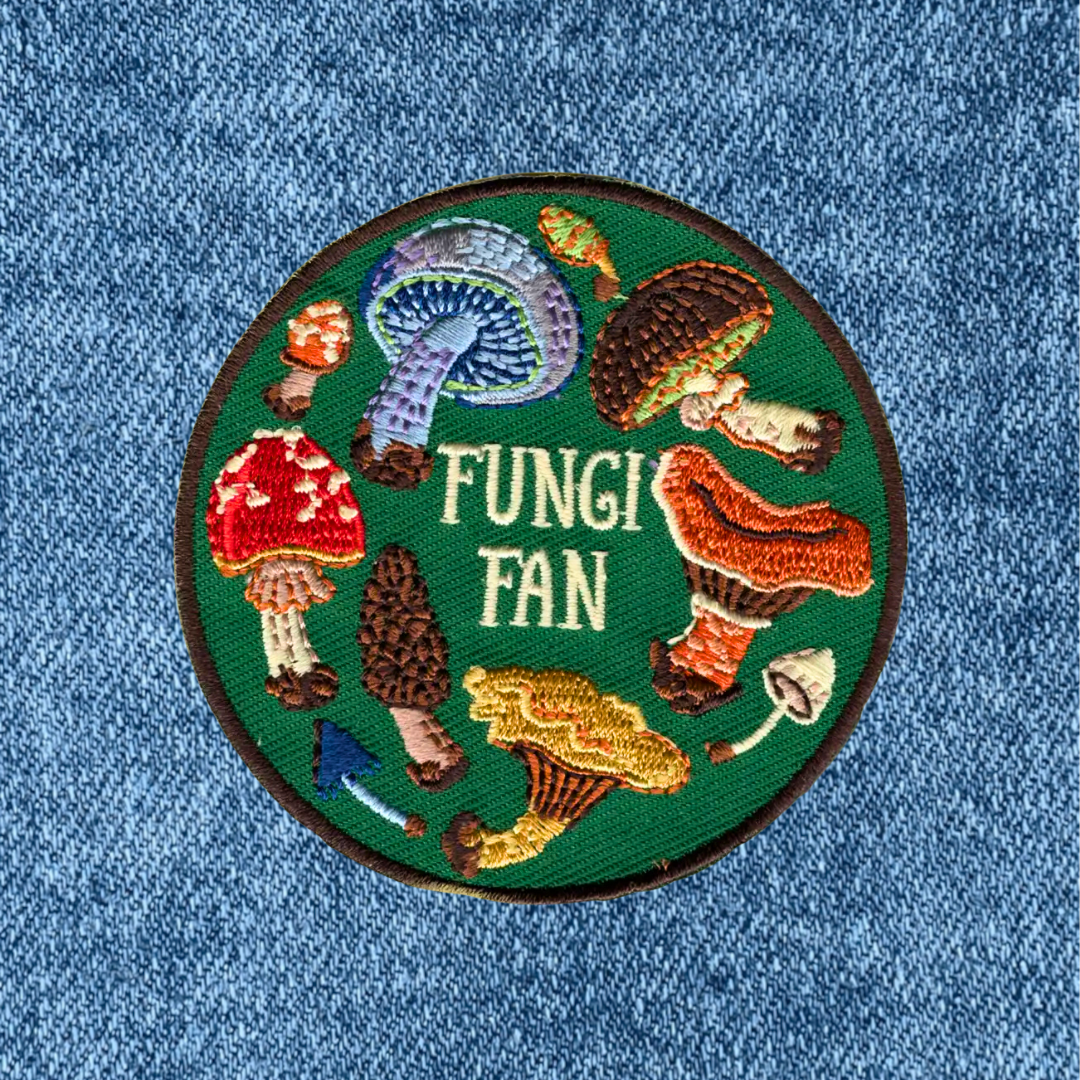 A circular embroidered patch with the words 'FUNGI FAN' in cream lettering, surrounded by various colorful embroidered mushrooms on a green background, placed on a blue denim fabric surface.