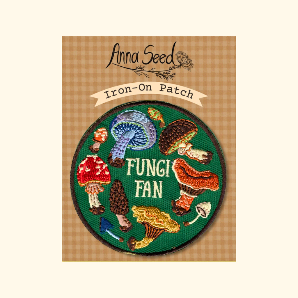 An embroidered iron-on patch featuring colorful mushrooms around the words 'FUNGI FAN' on a green background, presented on a tan gingham card labeled 'Anna Seed Iron-On Patch'.