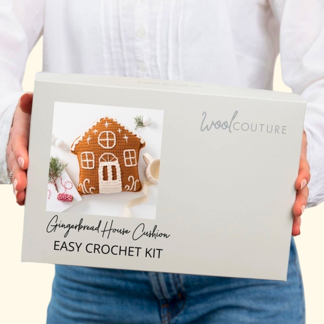 A person in a white blouse and blue jeans holds a large white box for a Wool Couture Gingerbread House Cushion Easy Crochet Kit. The front of the box features a photo of a crocheted gingerbread house and product text.