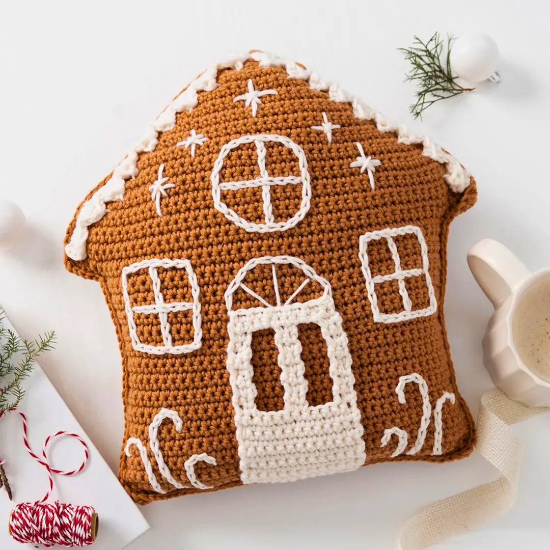 A crocheted gingerbread house pillow in warm brown with white yarn details, styled on a white surface with holiday props including a mug, ribbon, and pine sprigs.