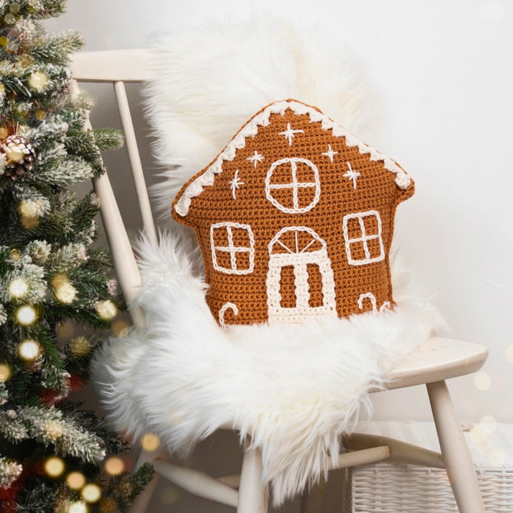 A crocheted gingerbread house pillow in warm brown and white, styled on a white faux fur cushion of a light wood chair next to a decorated Christmas tree.