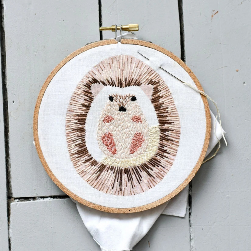Work in progress Kiriki Press Hedgehog Embroidery Kit displayed in a wooden embroidery hoop against a rustic grey timber background.