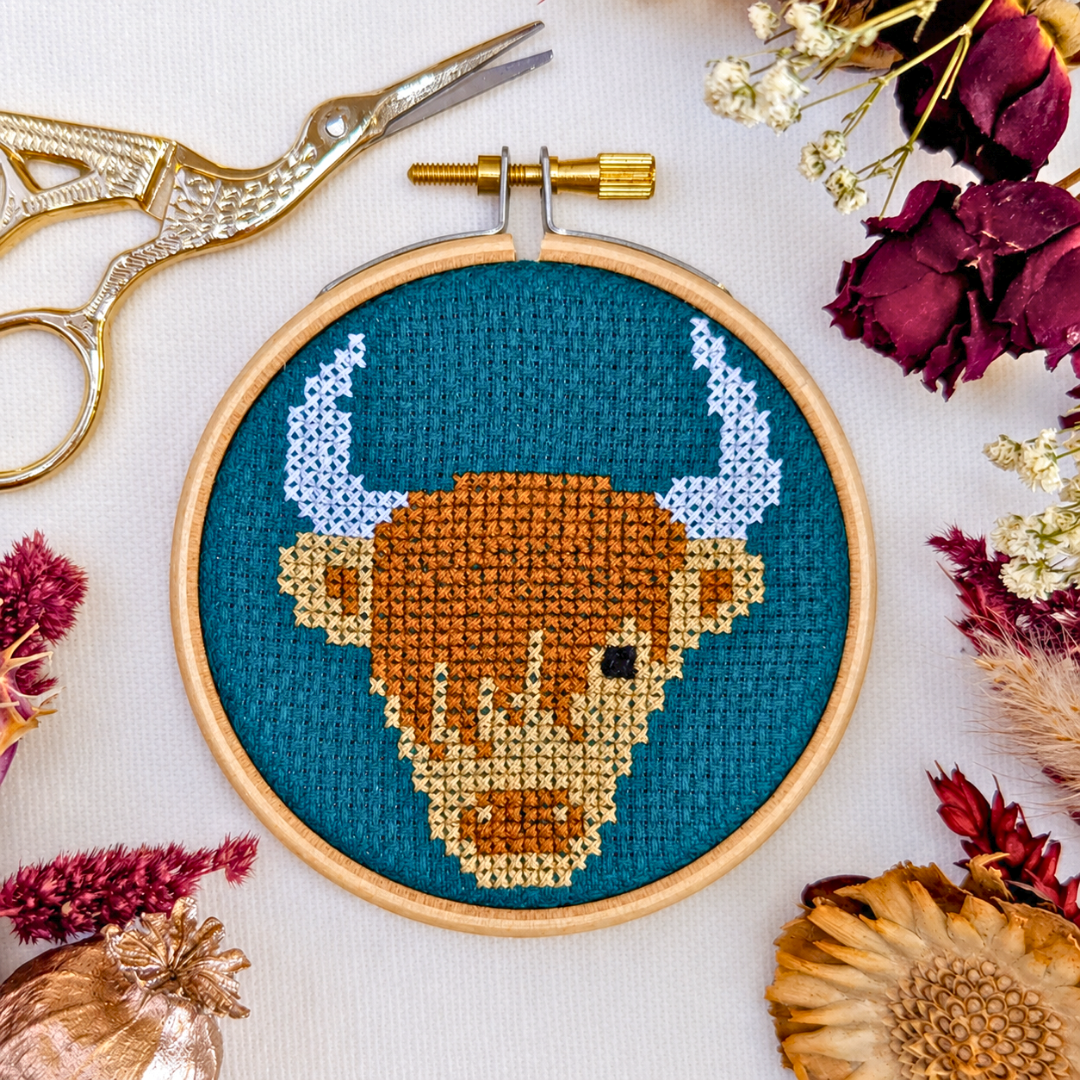 Finished Highland cow cross stitch displayed in a wooden hoop with flowers, leaves and craft scissors.