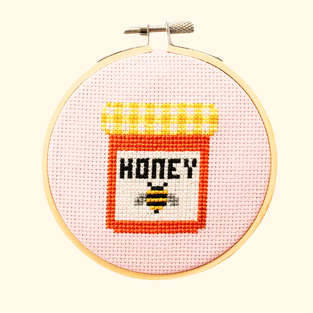 Finished honey jar cross stitch design in yellow, orange and black thread, displayed in a bamboo embroidery hoop on pink Aida fabric.