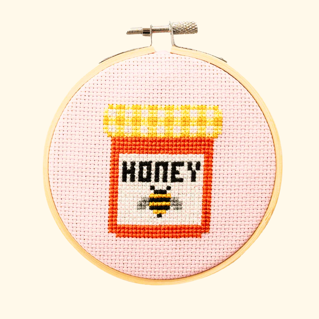 Finished honey jar cross stitch design in yellow, orange and black thread, displayed in a bamboo embroidery hoop on pink Aida fabric.